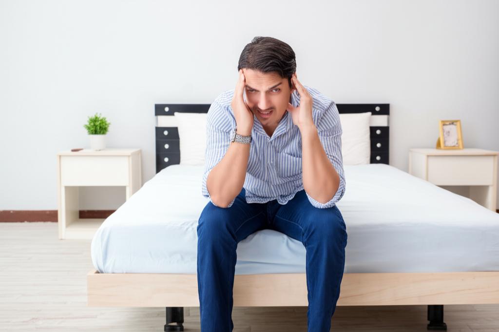 A middle-aged man looking stressed and frustrated while sitting on the edge of the bed.