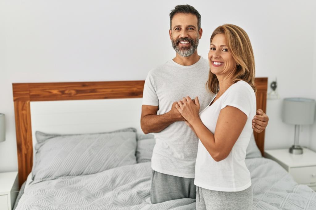 A happy middle-aged couple smiling and holding hands in their bedroom.